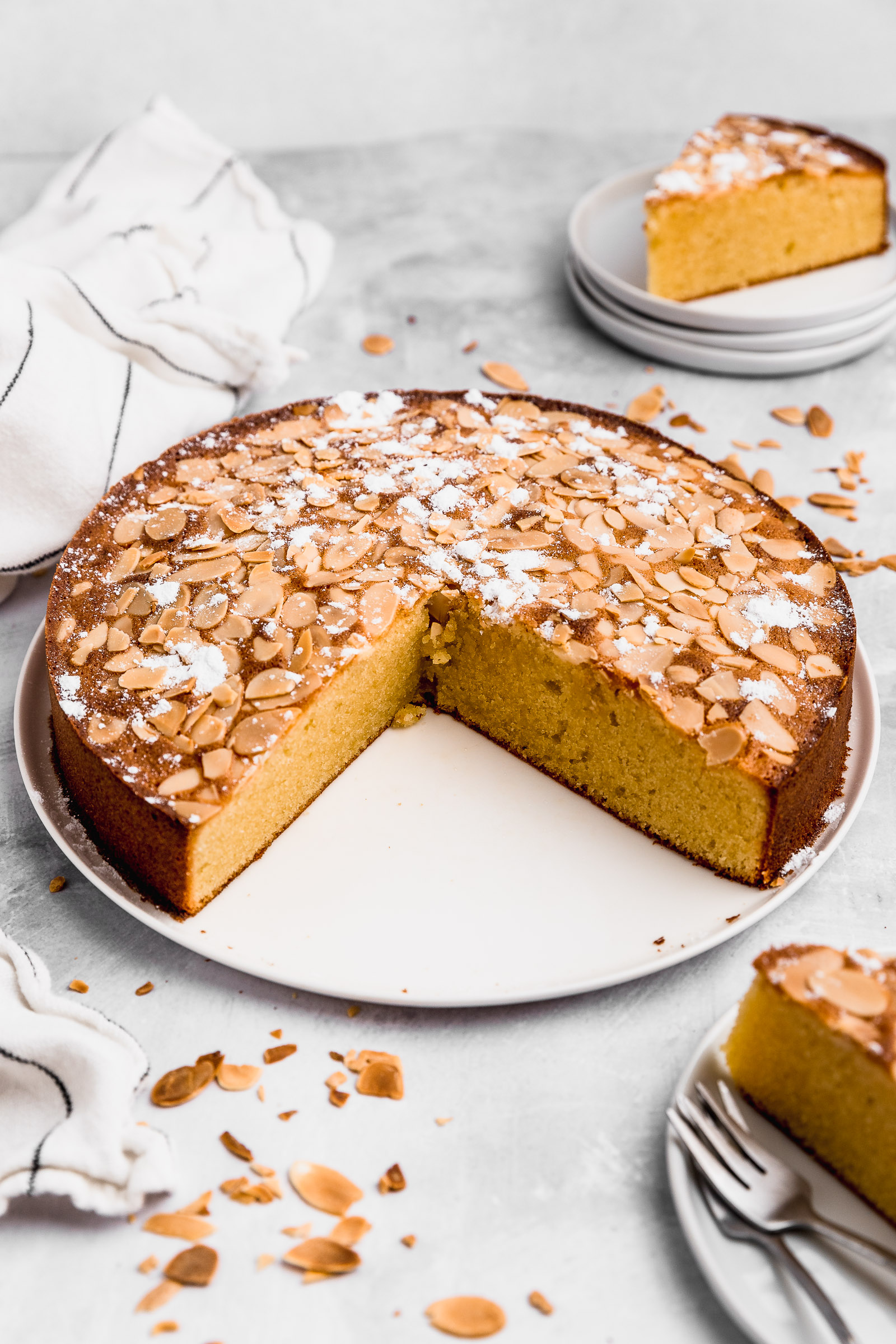 Front view of an almond cake (gluten-free) with toasted sliced almonds on top and icing sugar. Two pieces have been taken so that you can see the moist interior.