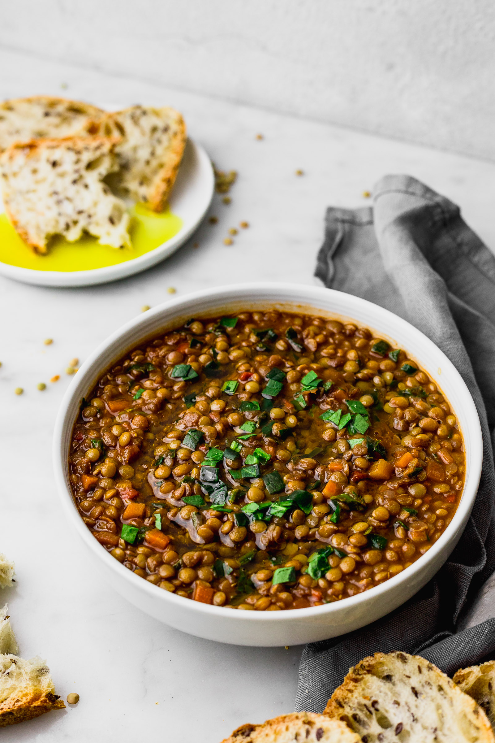 Brown lentils in a bowl, finished with olive oil and spinach. On the side, a small dish with olive oil and a slice of bread.