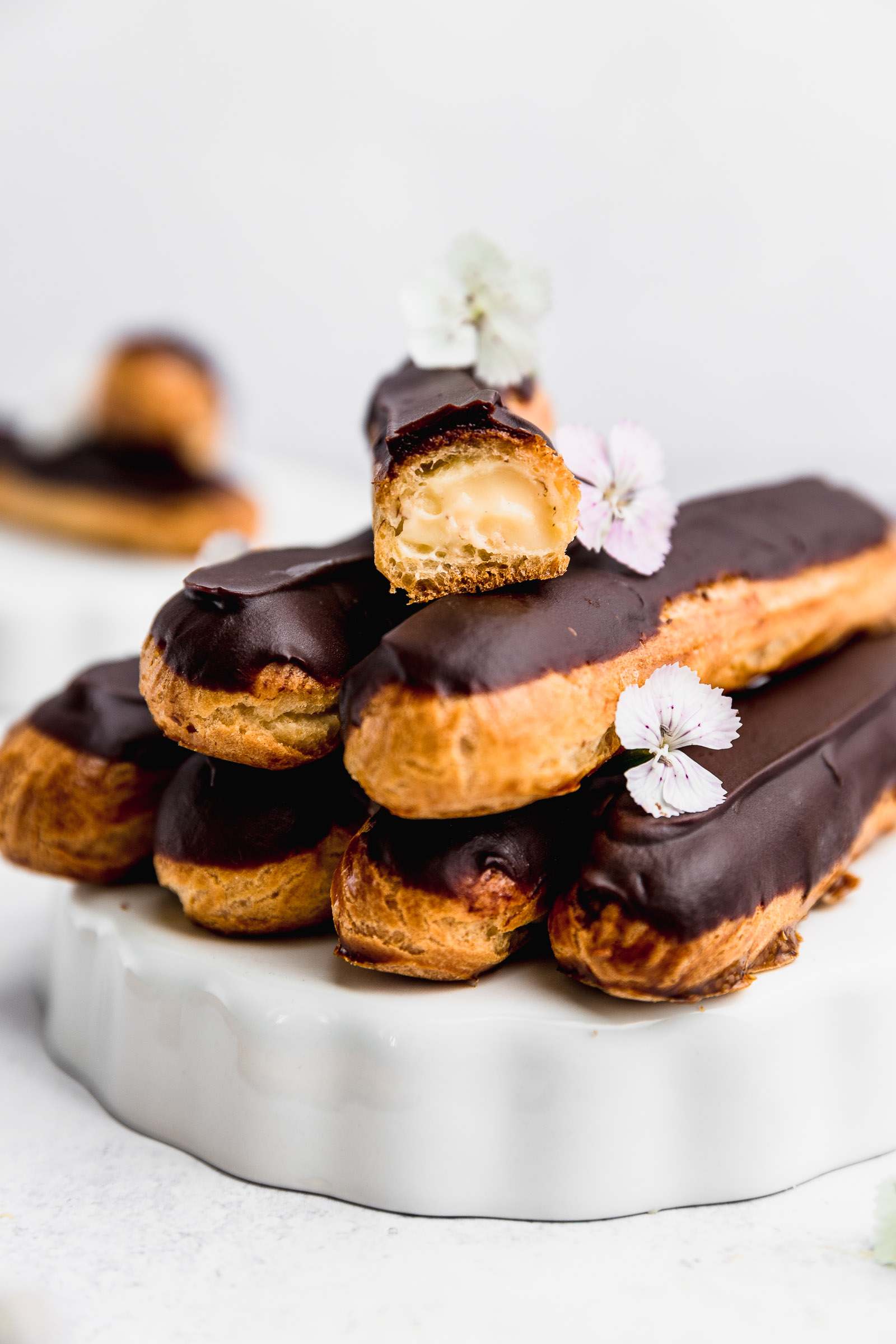 A pyramid of chocolate éclairs with for in the bottom, two on the second layer and one, split in half in a cross section to show the pastry cream on the inside. These are also light pink edible flowers laying on them.
