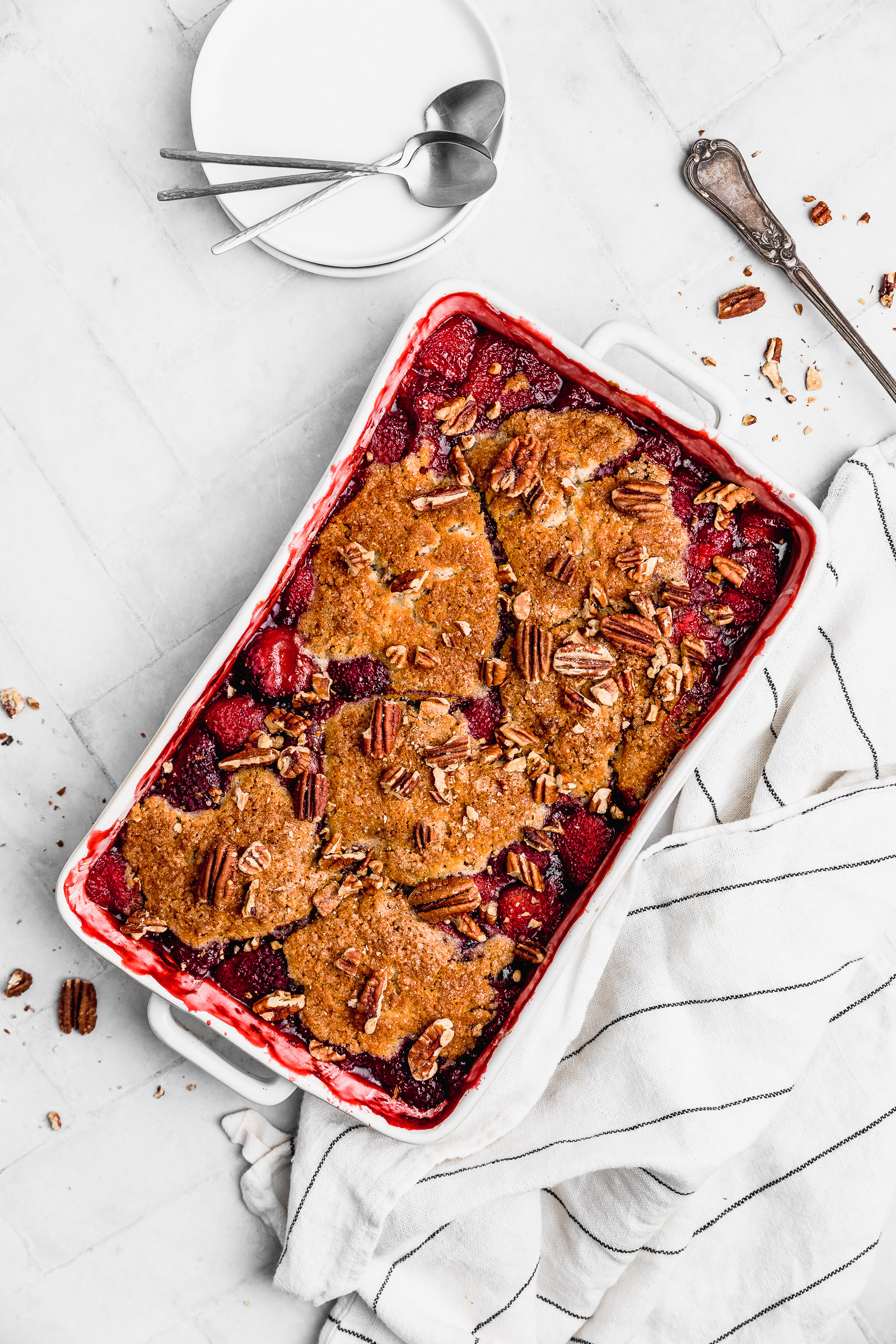Top view of a strawberry cobbler in a baking dish. It's crispy on top and you can see the filling peeking through as well. It's finished with chopped pecans.