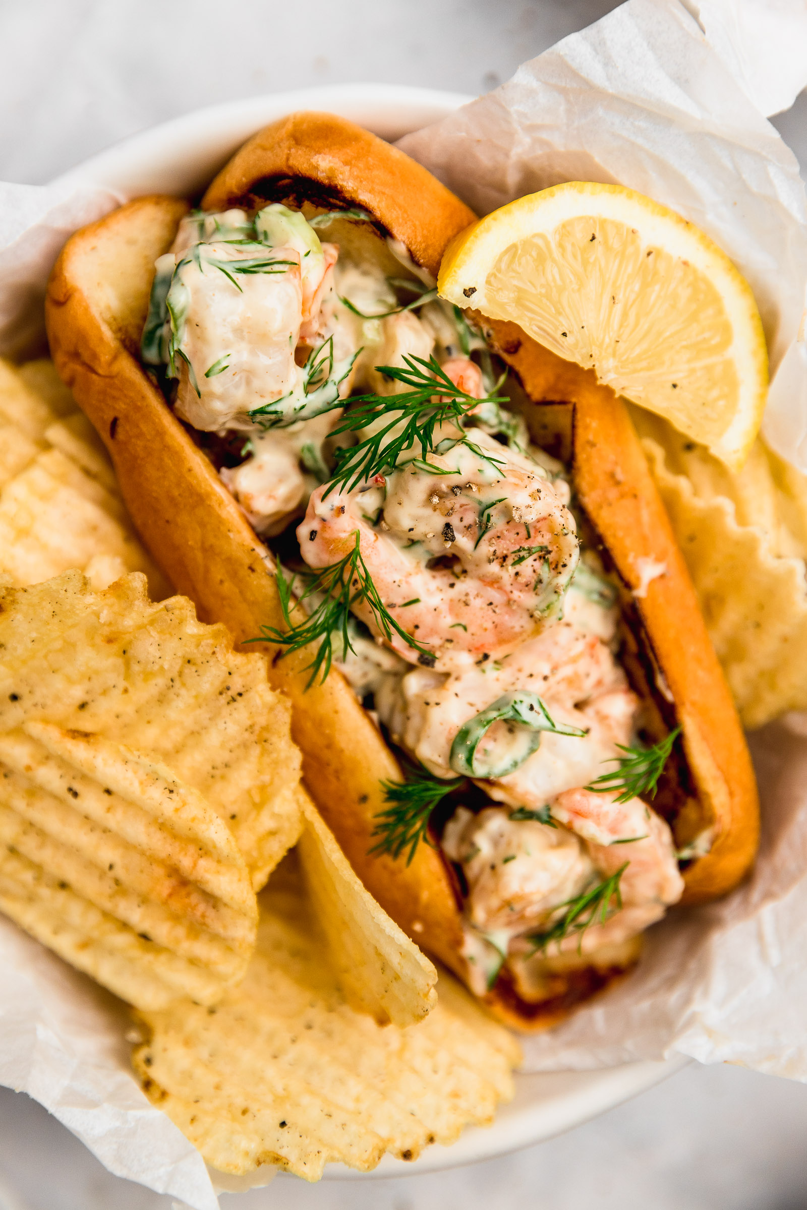 Closeup of a Shrimp Salad Sandwich in a hot dog bun, with extra dill on top and served with a side of chips and a lemon wedge.