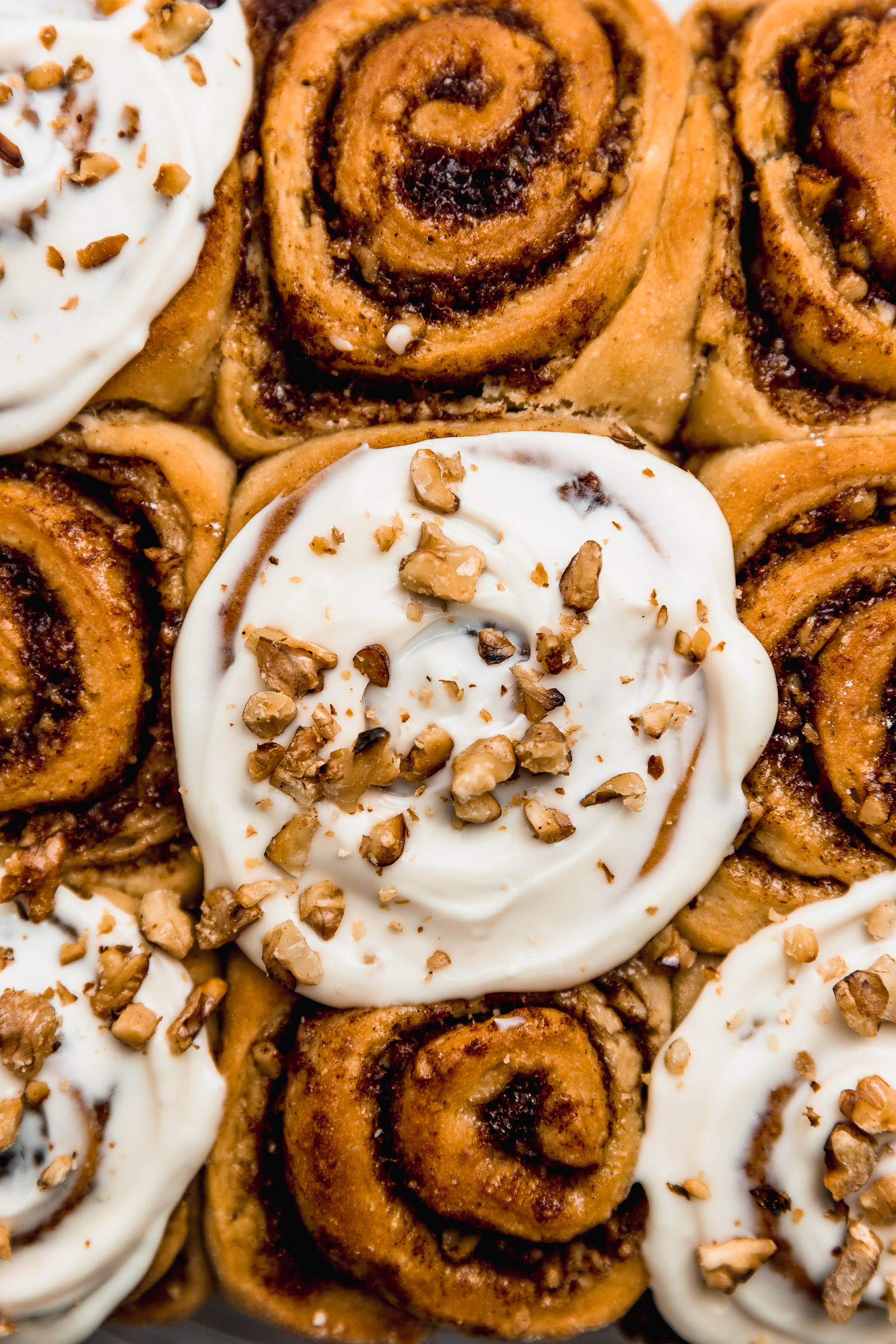 Closeup of banana cinnamon rolls. At the centre is one topped with cream cheese frosting and sprinkled with chopped walnuts. The ones on the side don't have frosting so you can see their shape and filling.