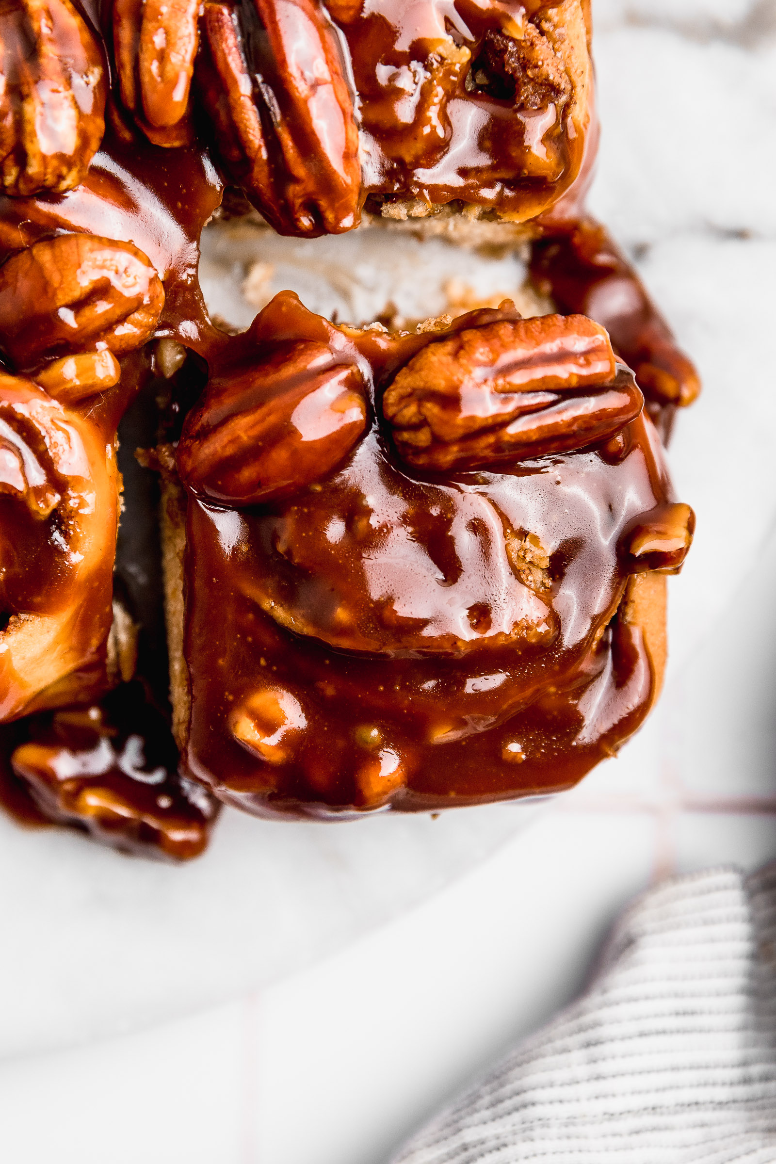 Closeup of a Caramel Pecan Cinnamon Roll, with that shiny sauce on top and you can see the whole pecans as well. It's being pulled apart from the rest of the sticky buns.