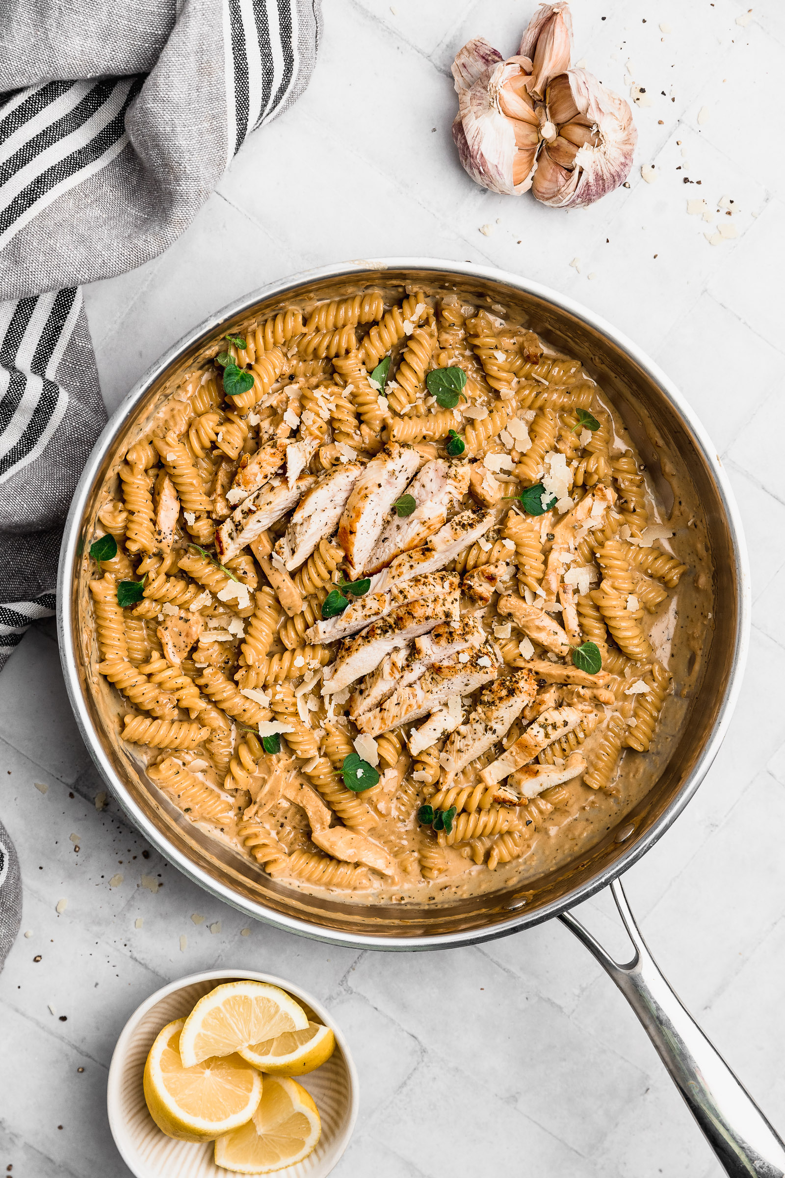 Top view of Garlic Parmesan Chicken Pasta contained in the pan where it was made. You can see the glossy and creamy sauce, as well as the golden chicken slices, fresh oregano leaves and parmesan flakes