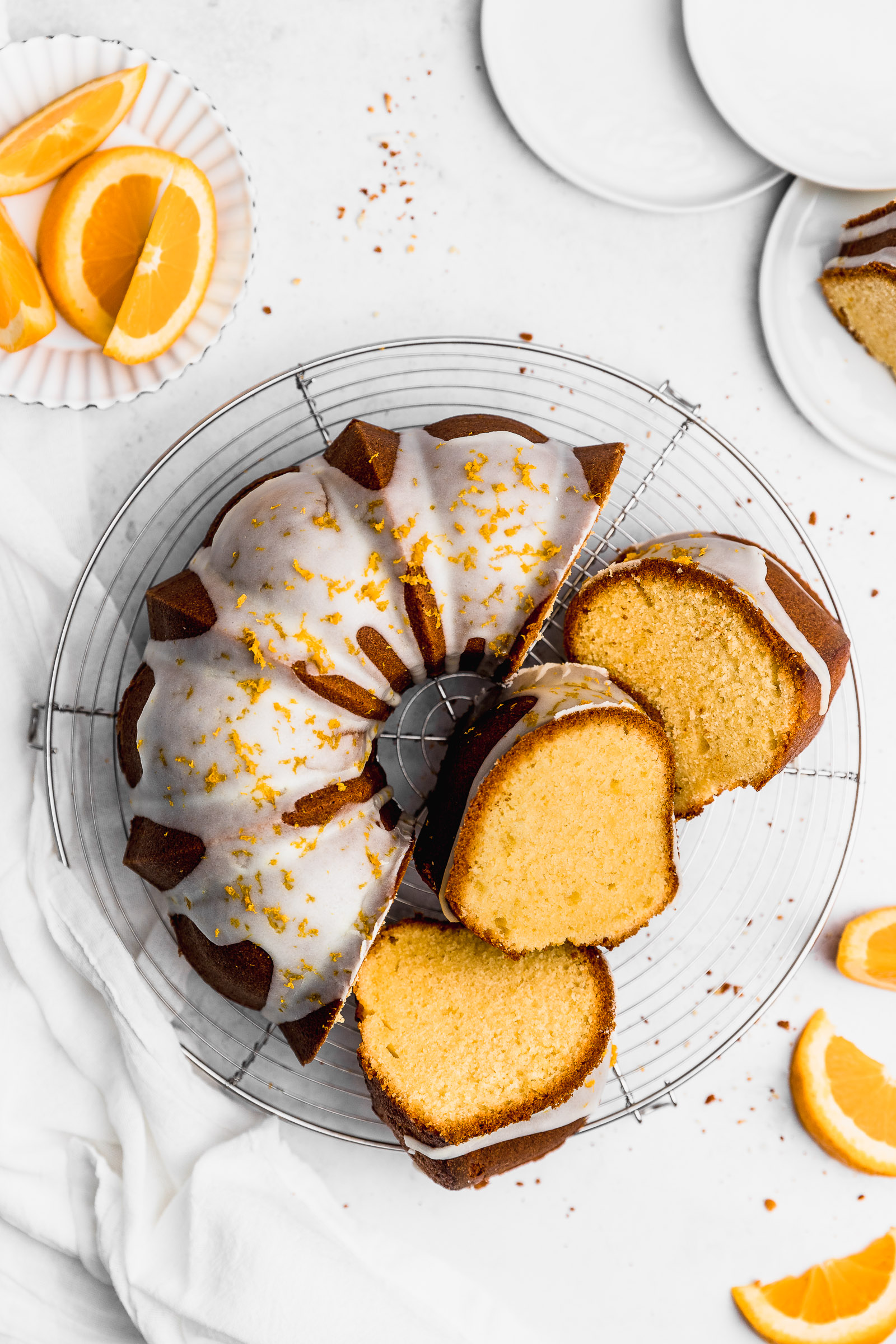 Top View of an Orange Bundt Cake, with a white glaze and orange zest on top. 3 slices lay on the side showing the crumb inside and that light orange colour.