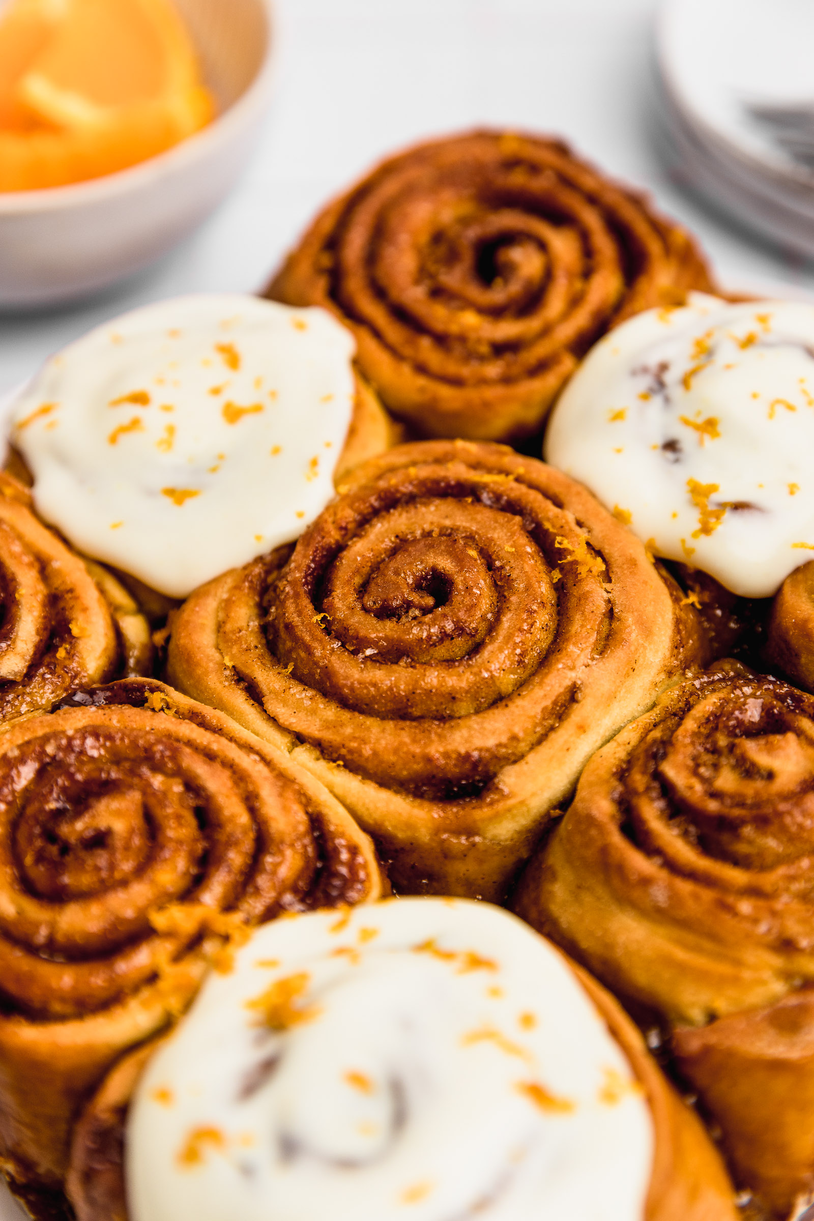 Closeup of orange cinnamon rolls, where you can see a clear roll and others in the background have orange cream cheese frosting on them.