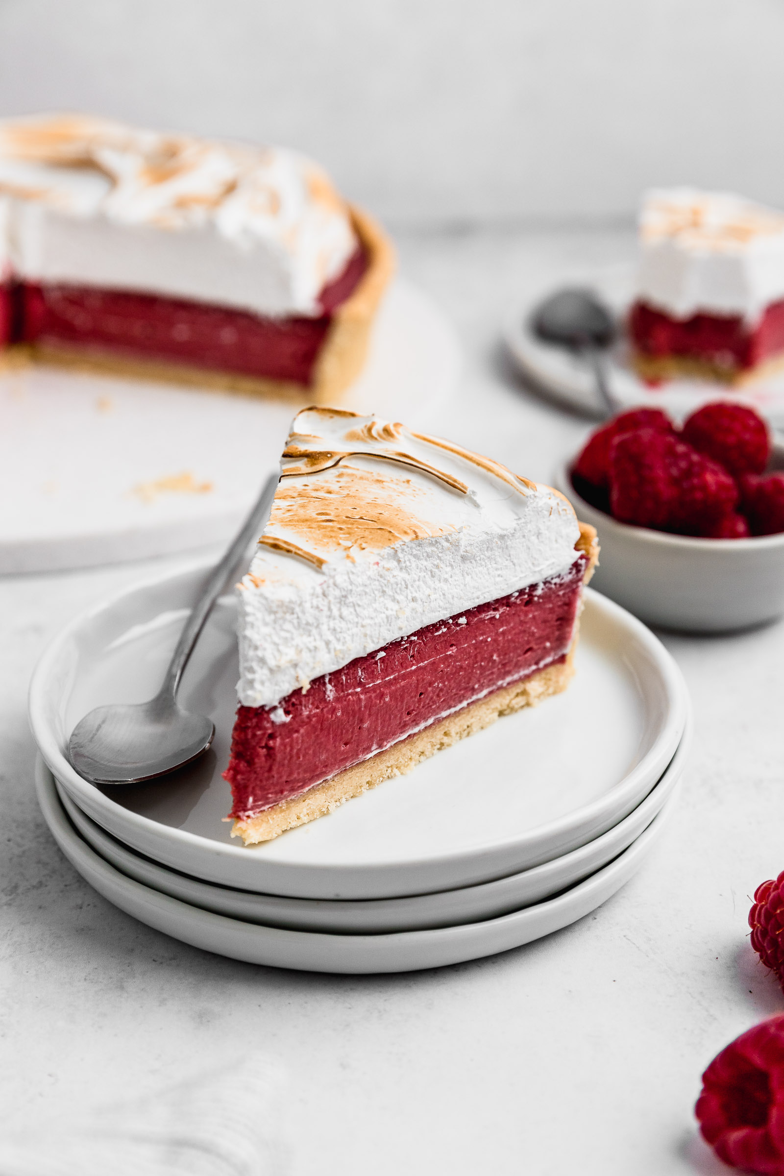 Slice of Raspberry Meringue Pie Recipe with Swiss Meringue. The layers are very evident with a golden crust, a bright red raspberry filling and a white Swiss Meringue on top that has been blow-torched.